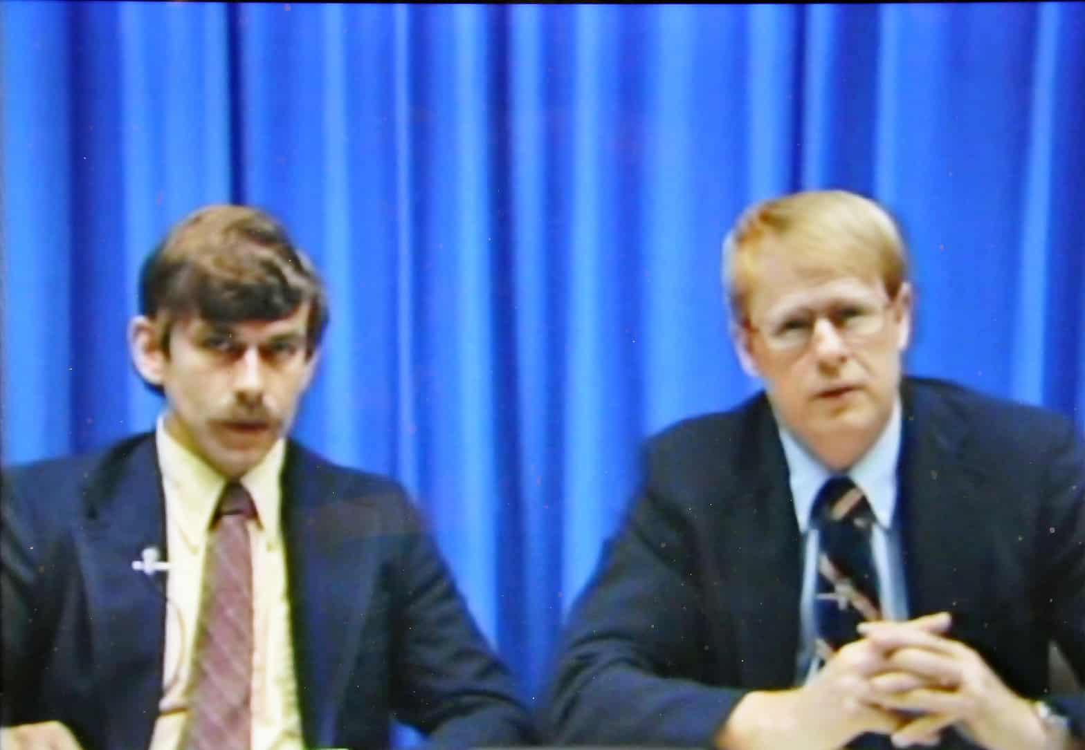 two men in suits sitting down giving a presentation