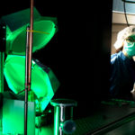 Two people in a dark space with reflected green and pink light. They wear hairnets and masks to keep the space clean