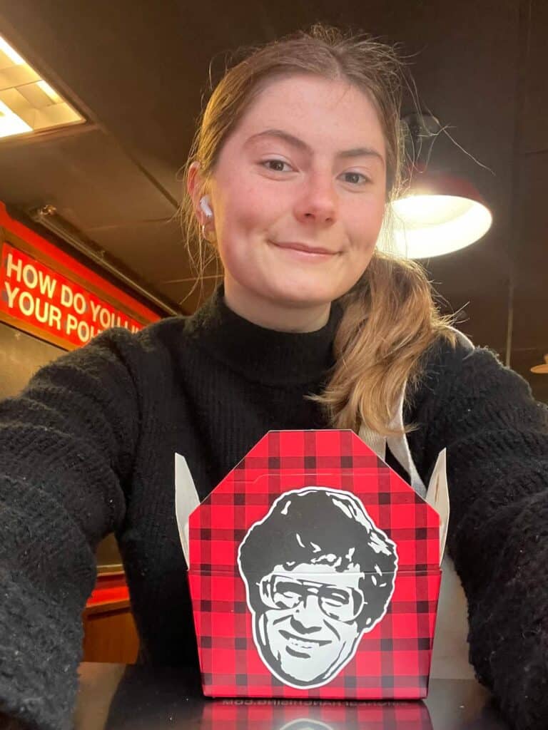 woman posing and smiling with a big ole' carton of poutine (the carton is red and black buffalo plaid)