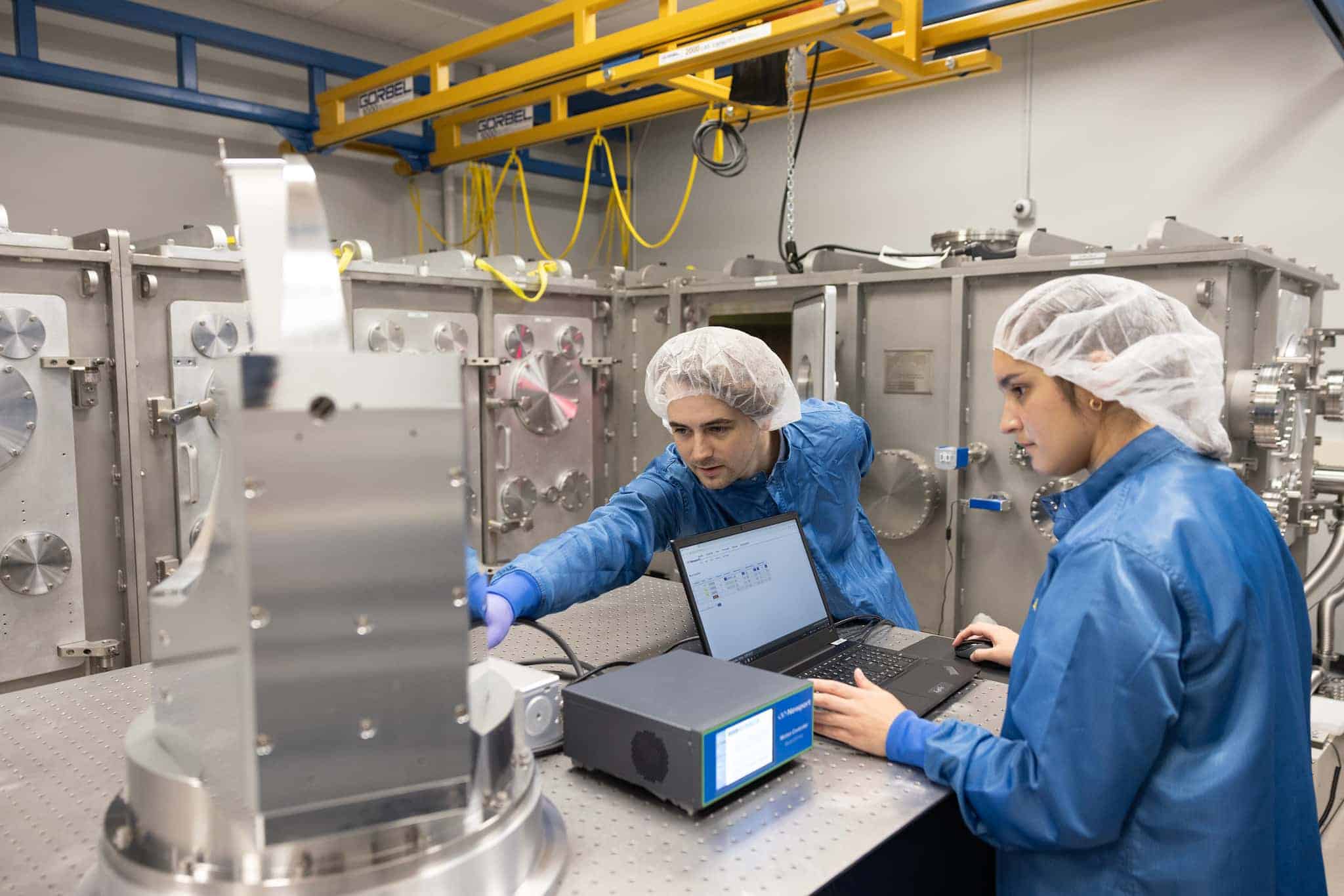 Two lab technicians in blue coats and hairnets work on a laptop next to a large metallic machine in a laboratory setting.