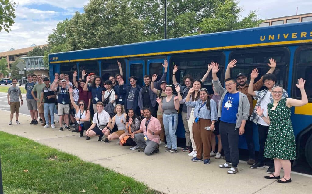 a group of students and faculty gather in front of a blue and maize bus