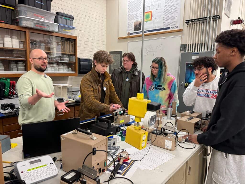 a faculty member speaks to a group of students gathered around equipment