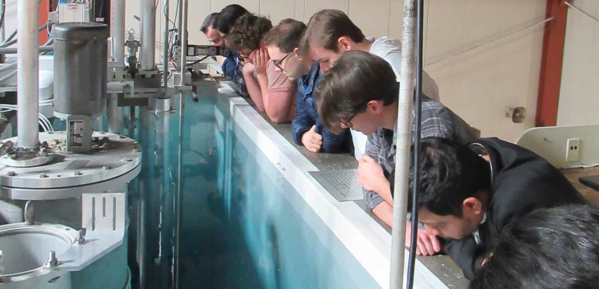 A group of people lean over the edge of a large pool filled with clear blue water, closely observing the contents below. The pool is part of a nuclear reactor or research facility, with metal machinery and instrumentation visible around and inside the water. The individuals, dressed in casual and lab-appropriate clothing, are engaged and focused on the activity or equipment within the pool.