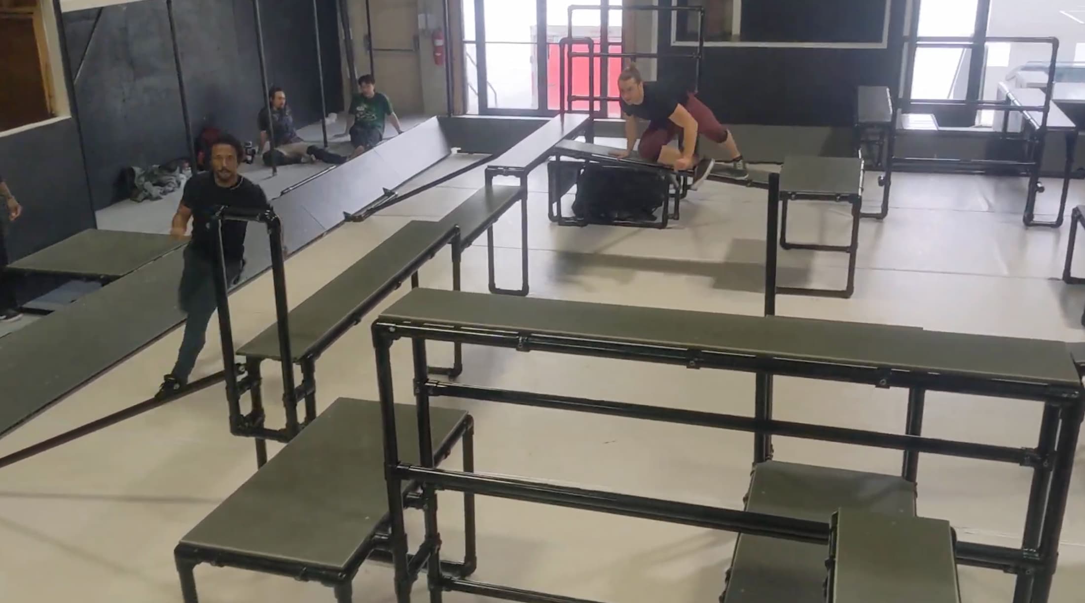 A facility with with bars, platforms, and obstacles. A man is running in one corner while another man leaps over an obstacle.