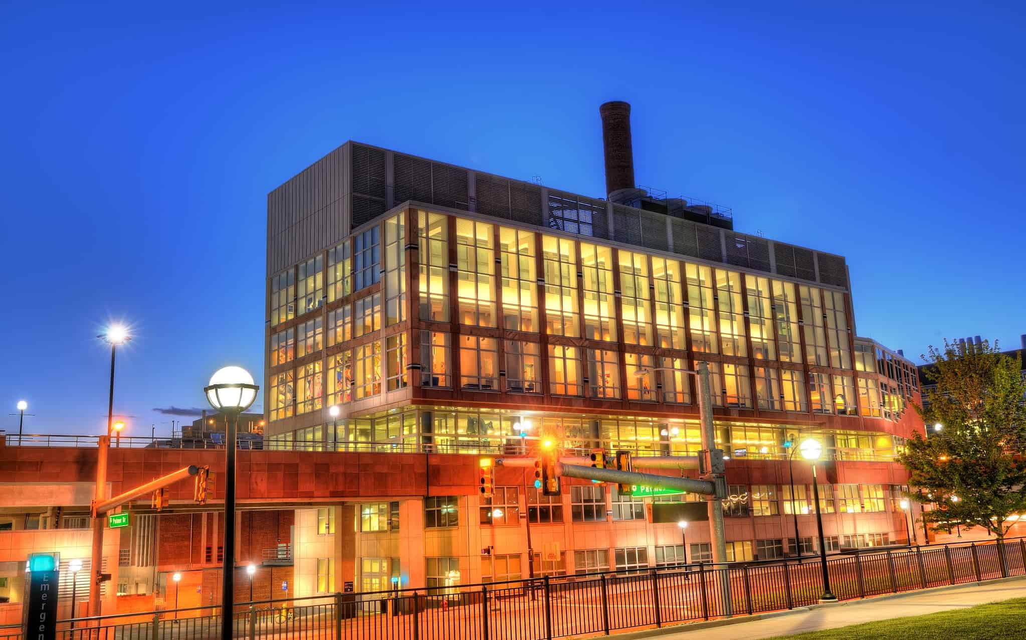 Palmer Commons at the University of Michigan, a modern glass-and-brick building illuminated at dusk, with its tall smokestack visible on the roof and vibrant lighting casting a warm glow against the evening sky.