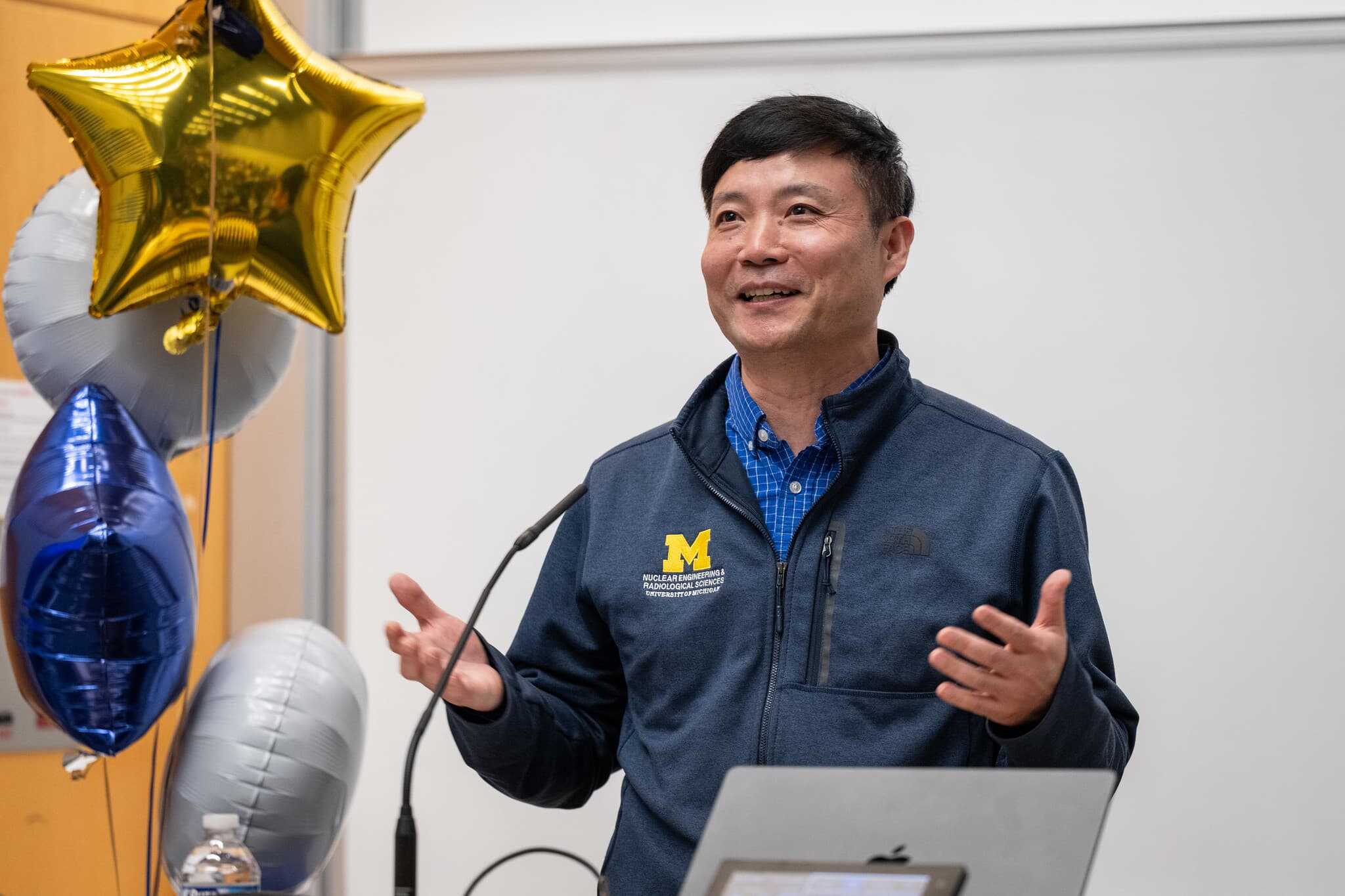 Zhijie (George) Jiao speaks at a podium during an award ceremony. He is wearing a University of Michigan Nuclear Engineering and Radiological Sciences jacket and smiling while gesturing with his hands. A cluster of metallic balloons, including a gold star, is visible beside him.