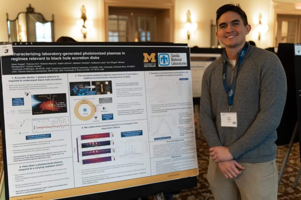 Isaac Huegel standing beside his research poster at a conference. The poster features diagrams and data on photoionized plasmas in black hole accretion disks, with logos for the University of Michigan and Sandia National Laboratories.