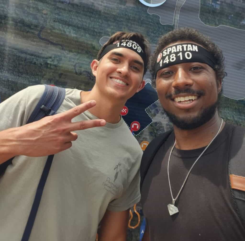 Isaac Huegel and a friend smiling after completing a Spartan Race, both wearing black race headbands and casual athletic gear, standing in front of a map of the course.