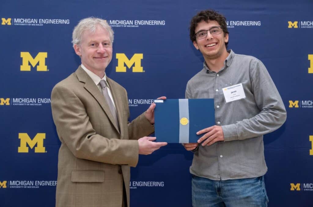 Jarett LeVan smiles while receiving the Distinguished Undergraduate Achievement Award from a Michigan Engineering faculty member. Both are standing in front of a University of Michigan Engineering step-and-repeat banner, and Jarett holds a blue award folder.
