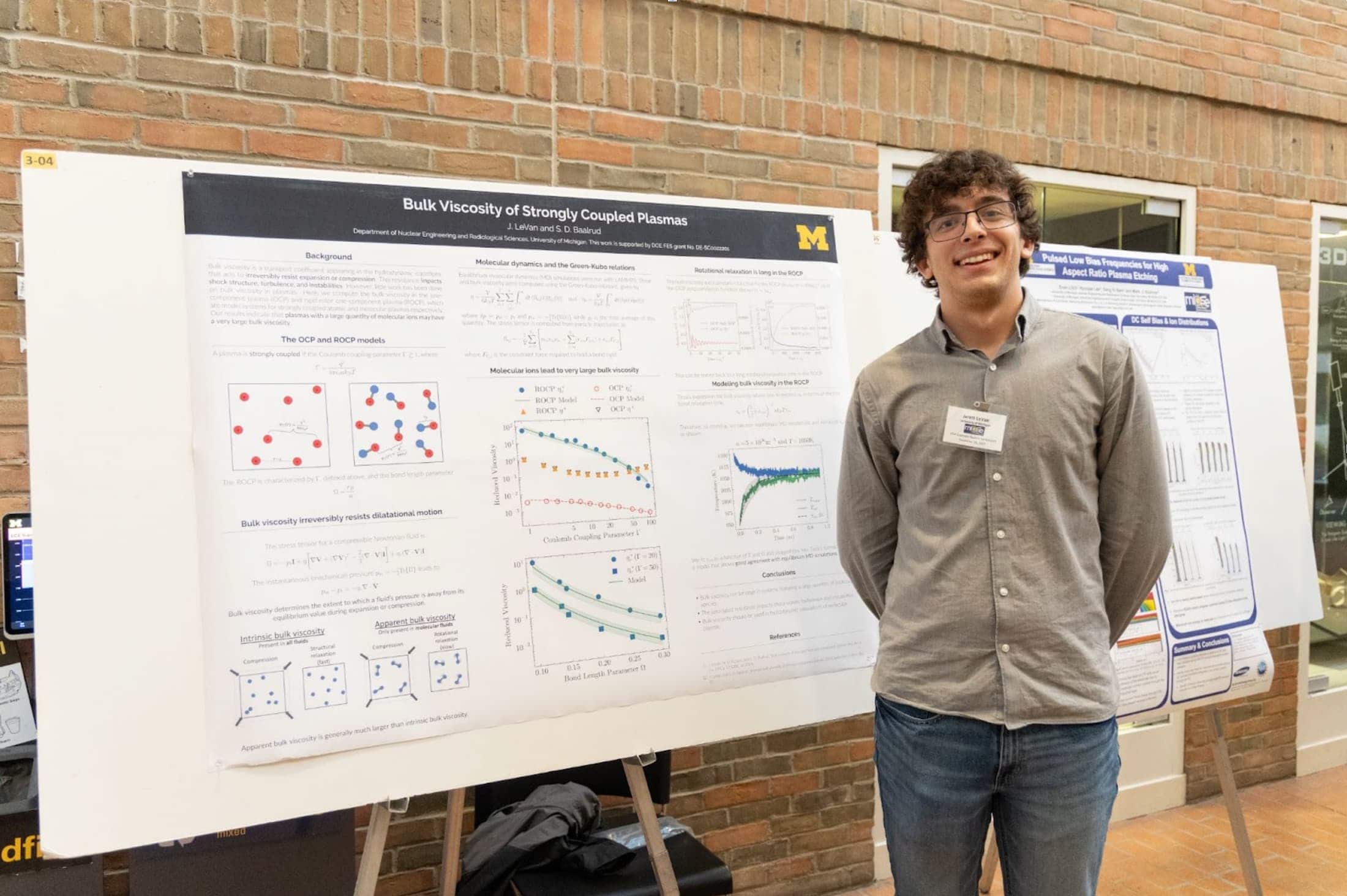 Jarett LeVan stands in front of his research poster titled "Bulk Viscosity of Strongly Coupled Plasmas" at a poster session. He is smiling and wearing a gray button-down shirt and jeans, with a brick wall and other posters in the background.