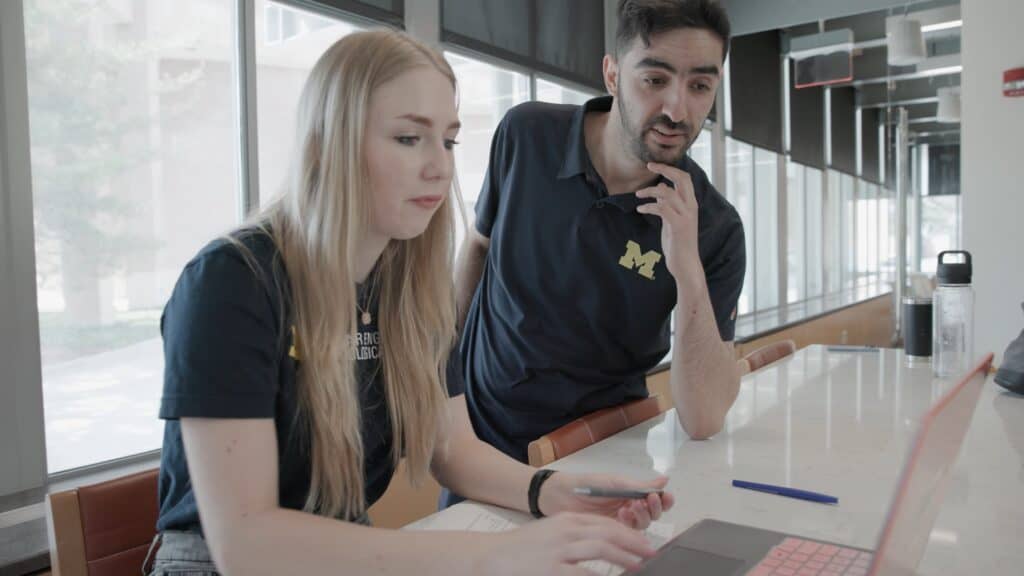 A University of Michigan professor and a student collaborate at a table near large windows. The student is seated, typing on a laptop and holding a pen, while the professor leans over to look at the screen. Both wear dark blue shirts with maize block M logos. Papers, a pen, and water bottles are visible on the table.