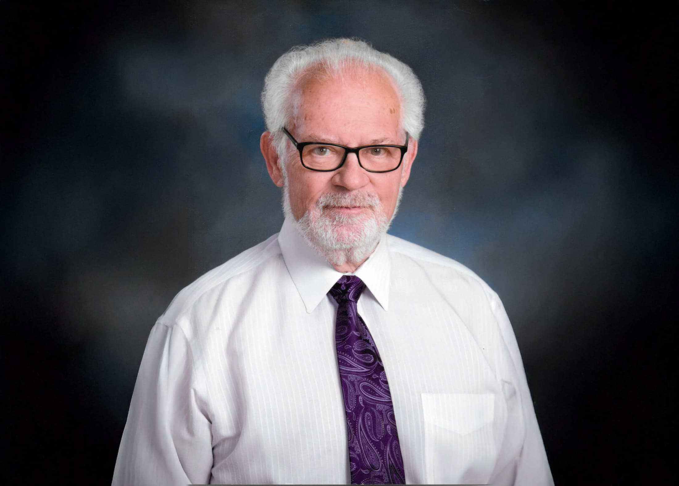 Man with white hair and a beard wearing black glasses, a white dress shirt, and a purple paisley tie, posed against a dark studio background.