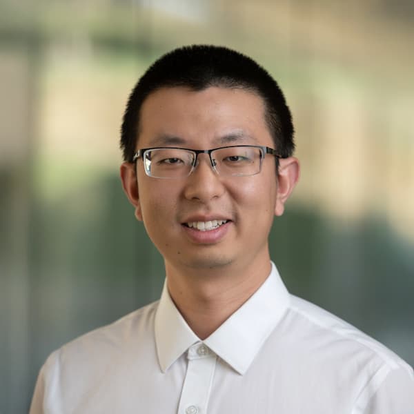 A professional headshot of Shuai Che, who is wearing glasses and a white collared shirt. He is smiling and standing in front of a softly blurred background.
