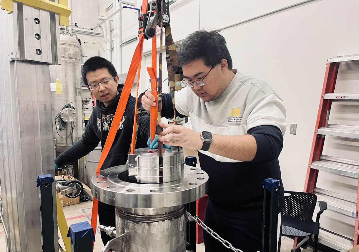 Shuai Che (left) and Muyue Li (right) are installing the bushing-type mechanical seal on the Shaft Seal Test Facility. Che wears a Star Wars hoodie, and Li wears a Michigan Engineering sweatshirt while working with orange lifting straps and a metal seal component. The setup includes a chain-secured test stand and lab equipment in the background.