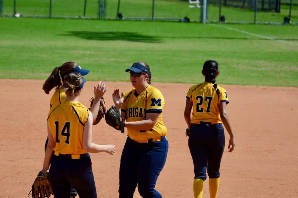 Colleen Campbell (#24) high-fives teammates on the field during a game. Wearing yellow jerseys and navy pants, the Michigan players gather on the dirt infield after a successful play.