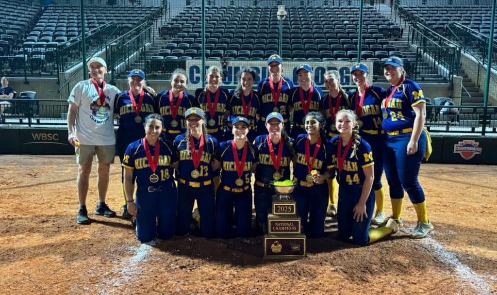 The University of Michigan club softball team poses on the field after winning the 2025 national championship. All players wear navy and maize uniforms with gold medals around their necks, gathered around a large trophy labeled “2025 National Champions.”