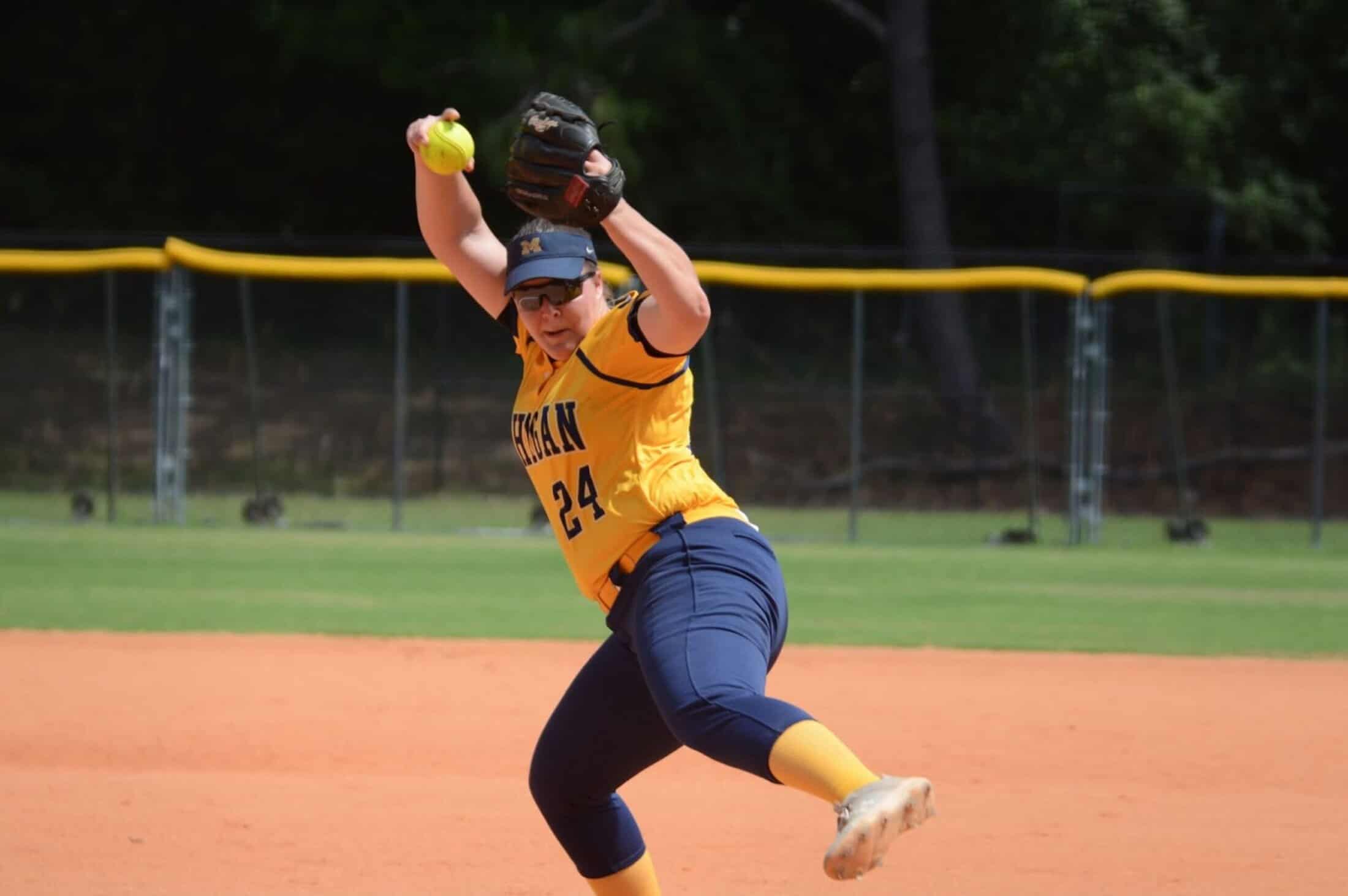 Colleen Campbell (#24) winds up mid-pitch, captured in motion with her arm raised and one foot off the ground. She wears a yellow Michigan jersey, navy pants, and a navy visor, focused on delivering the softball.