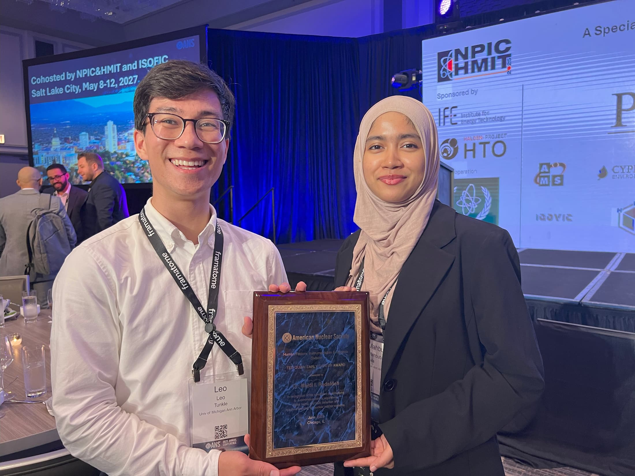 Two smiling individuals stand in front of a stage at a professional conference. The person on the left, wearing glasses and a white shirt, has a conference badge that reads "Leo, Univ of Michigan Ann Arbor." The person on the right, wearing a light pink hijab and black blazer, holds a wooden plaque with a blue background. Behind them, a screen displays the event name: “NPIC&HMIT and ISOFIC, Salt Lake City, May 8–12, 2027.”