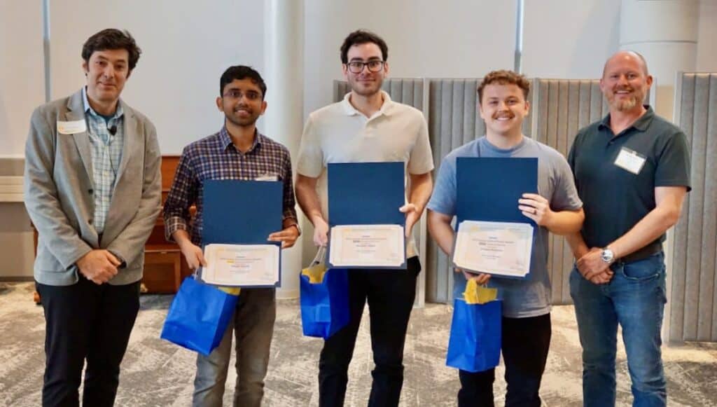 Five individuals stand in a row at an award ceremony. Three in the center hold certificates and blue gift bags. Crewse Petersen, wearing a gray T-shirt and holding his award, stands second from the right.