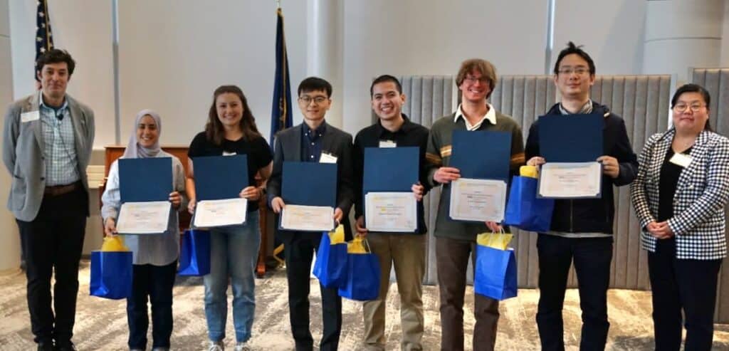 A group of eight people stands in a row at an award ceremony. Six individuals in the center hold certificates and blue gift bags with yellow tissue paper. Lucas Trojanowski, wearing a green sweater and glasses, is third from the right.