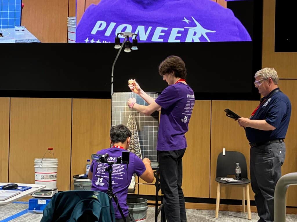 Two students in matching purple "PIONEER" shirts test a wooden tower structure at a Science Olympiad event, while a judge observes and takes notes. Equipment and buckets are set up around them in a modern auditorium.