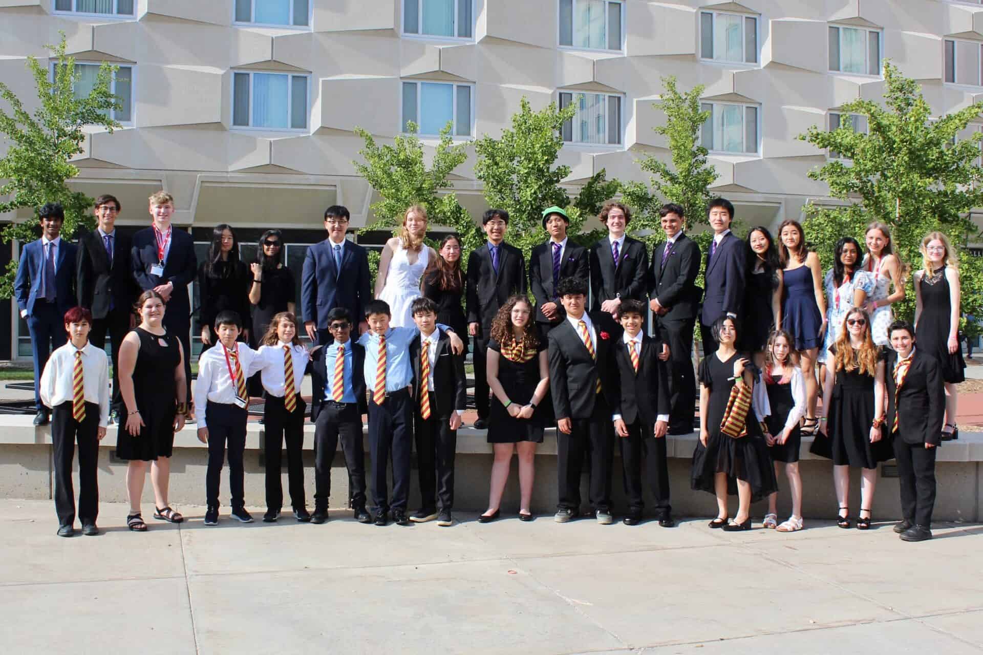 A large group of middle and high school students dressed in formal and semiformal attire pose outside a building with geometric windows, some wearing medals and matching ties, representing their Science Olympiad team.