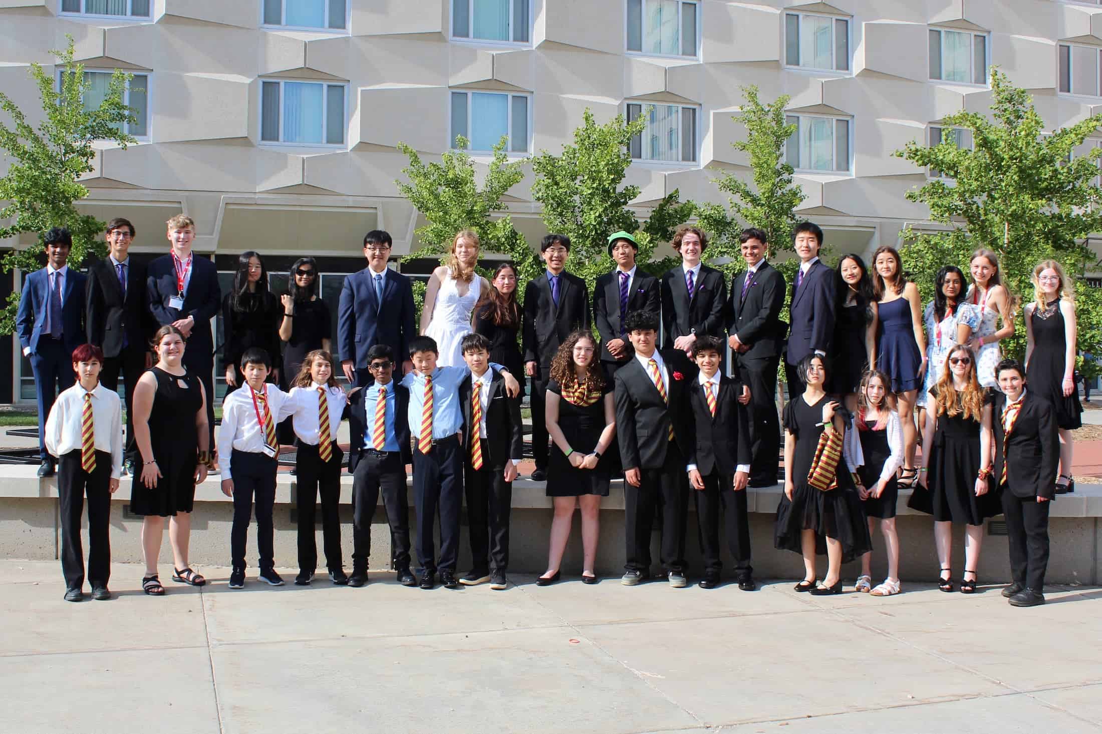 A large group of middle and high school students dressed in formal and semiformal attire pose outside a building with geometric windows, some wearing medals and matching ties, representing their Science Olympiad team.