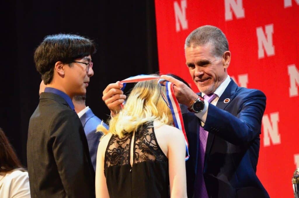 A man in a suit places a red, white, and blue medal around a female student's neck during an award ceremony, while another student looks on. The red backdrop features large white "N" letters for the University of Nebraska.