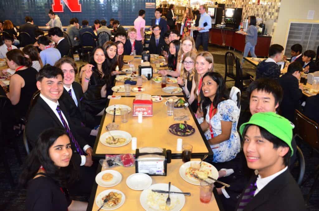 Science Olympiad team members dressed in suits and dresses smile around a long table during a group meal in a university dining hall. Plates of food and dessert are scattered on the table as students pose for the photo.