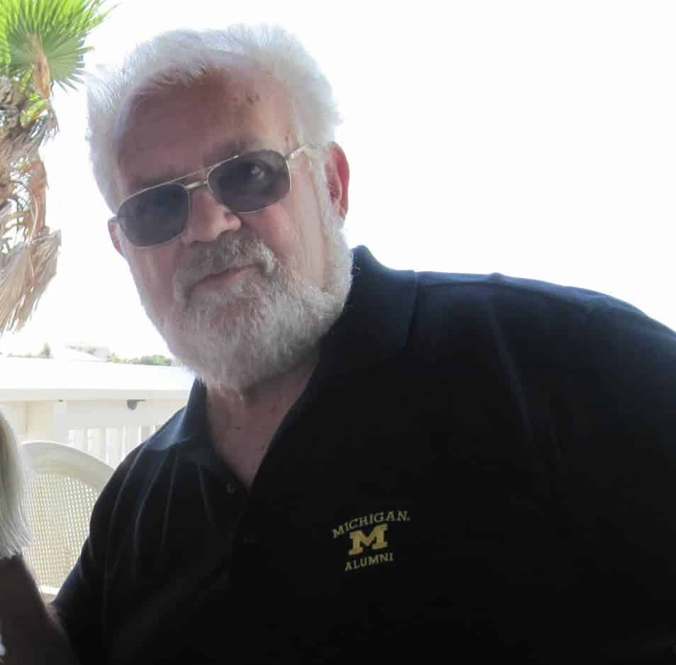 Man with white hair and a beard wearing sunglasses and a black “Michigan Alumni” polo shirt, standing outdoors near a palm tree on a sunny day.