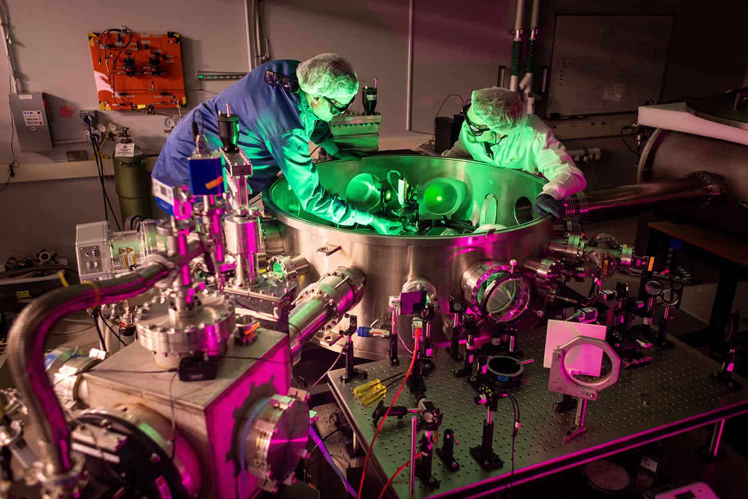 Two researchers wearing lab coats, hairnets, gloves, and protective glasses work inside a laser laboratory, adjusting components within a large, circular stainless steel vacuum chamber. The lab is illuminated with vibrant green and magenta lighting, highlighting the complex network of lasers, optics, and instrumentation surrounding the chamber.