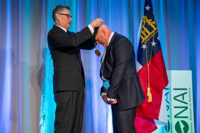Clint Ballinger receives a medal during the National Academy of Inventors 2024 Fellows Induction Ceremony. They stand in front of a blue curtain with the Georgia state flag and an NAI banner.
