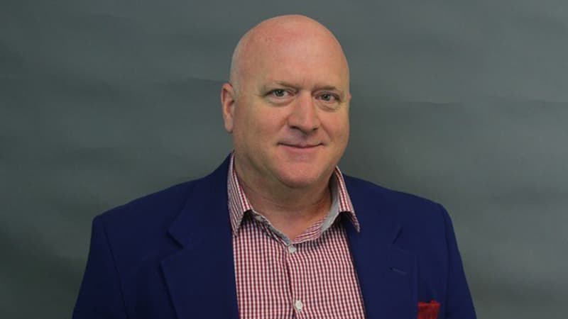 Clint Ballinger poses in front of a gray background wearing a navy blazer and red checkered shirt, smiling at the camera.